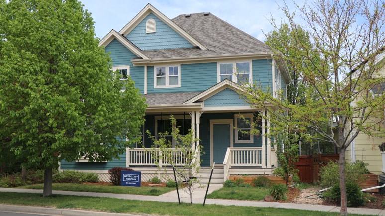 Green, Victorian style home in the Central Park neighborhood of Denver, Colorado with blue Metro City Roofing sign in the foreground