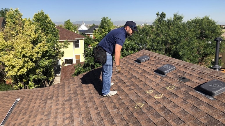 Metro City Roofing salesperson in a blue shirt and black hat inspecting a red shingle roof and marking hail impact marks in yellow chalk in Denver, Colorado with blue sky in the background.